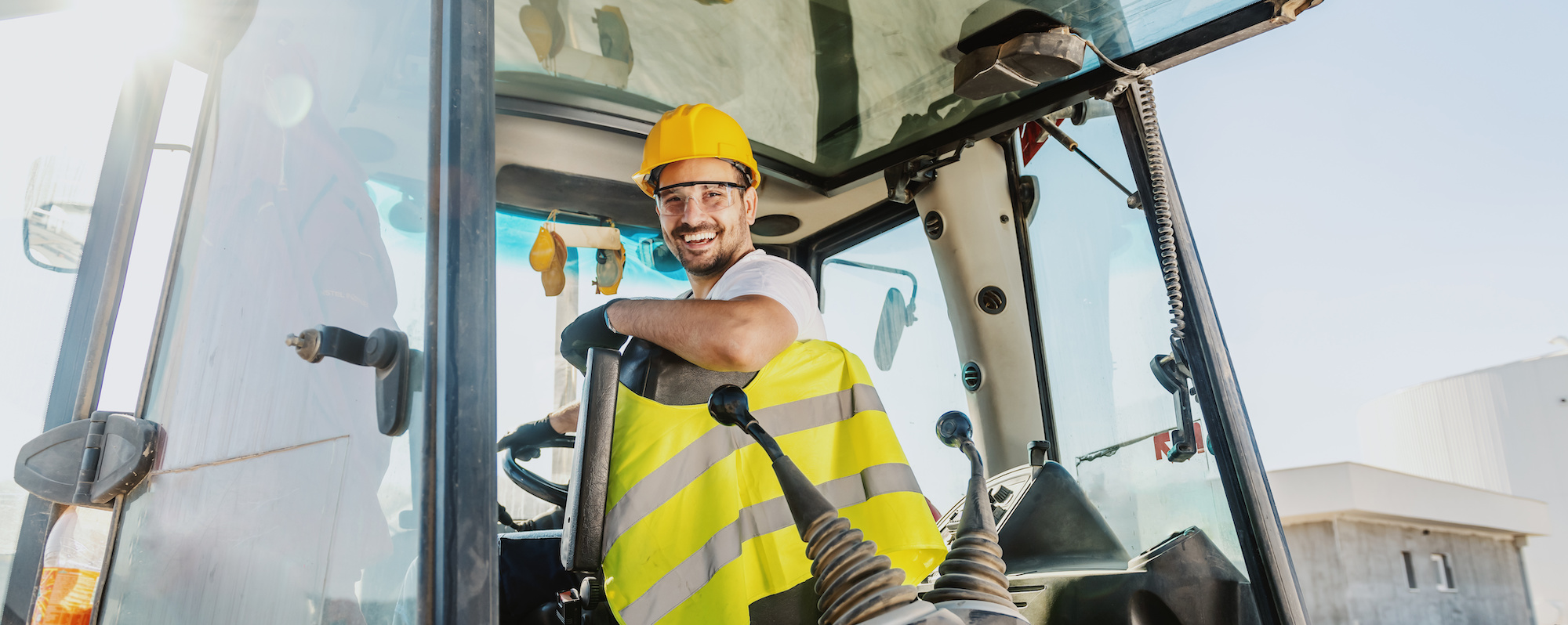 Smiling handsome caucasian worker in overall and with helmet on head driving excavator.